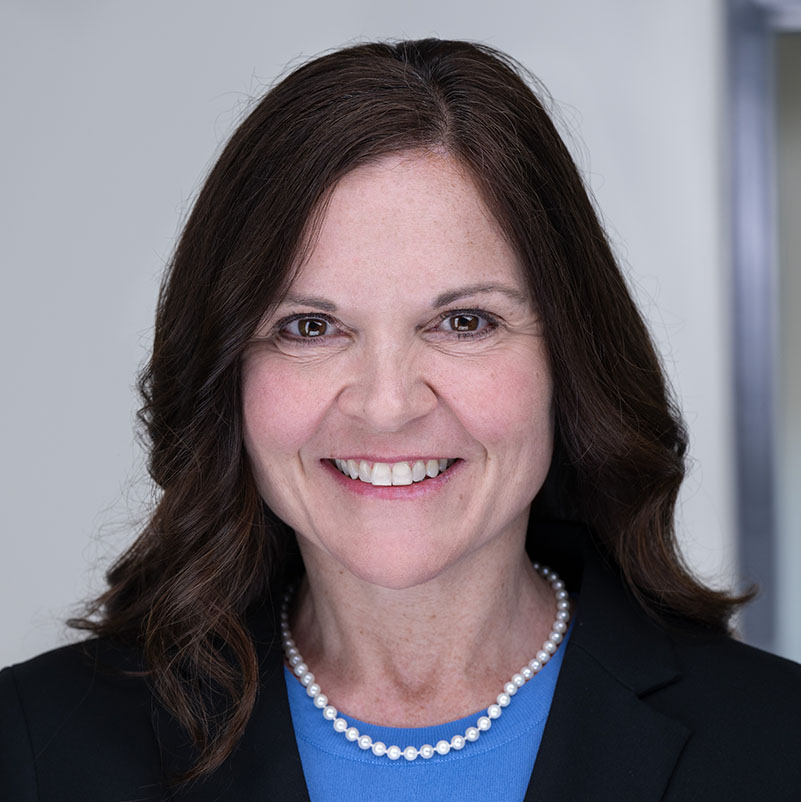 Headshot of Rebecca Coyle, with wavy long brown hair, a black blazer, string of pearls, and a royal blue shirt.