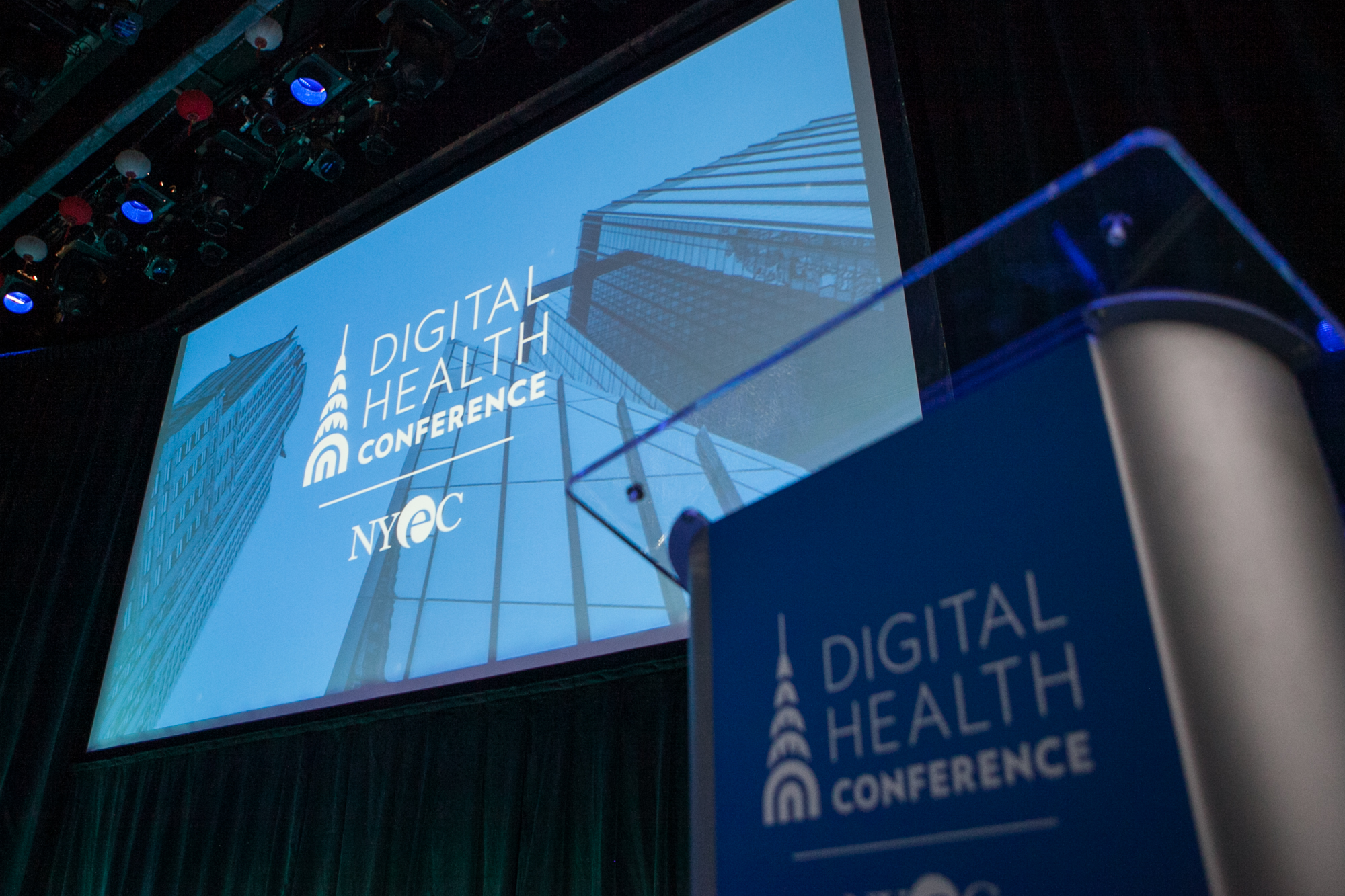 A large projector screen hangs beneath stage lights, with a promotional graphic cast onto it that reads Digital Health Conference N Y E C. A podium made from metal and glass sits in the foreground.